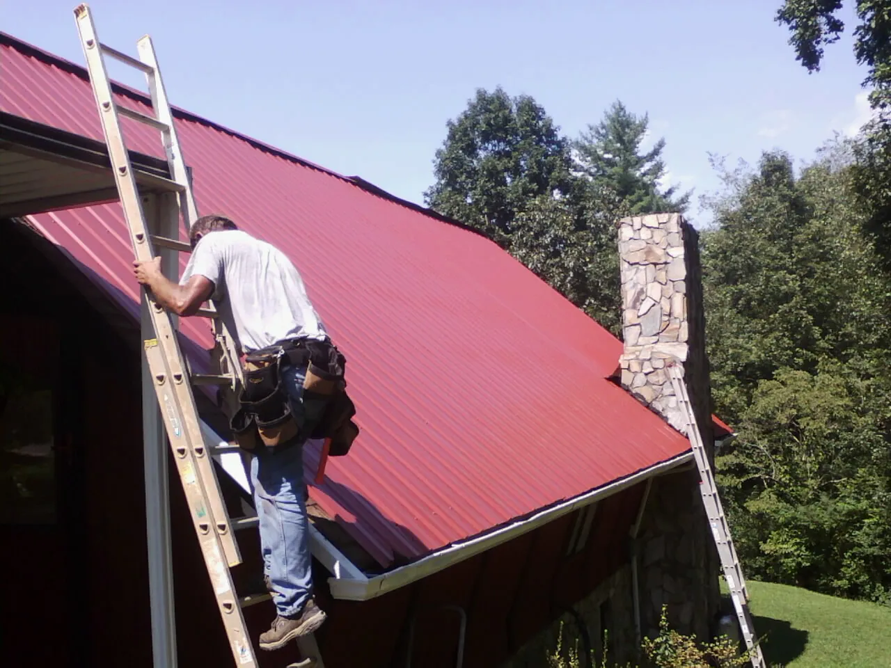 Roofer on a ladder beside a red metal roof and stone chimney — Soffit & Fascia Repair by Roofing Brothers in Skokie